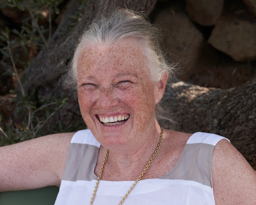 Close-up of an elderly woman with gray hair laughing outdoors, wearing a white and beige dress and a gold chain necklace.