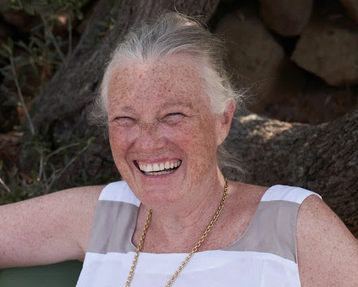 Close-up of an elderly woman with gray hair laughing outdoors, wearing a white and beige dress and a gold chain necklace.