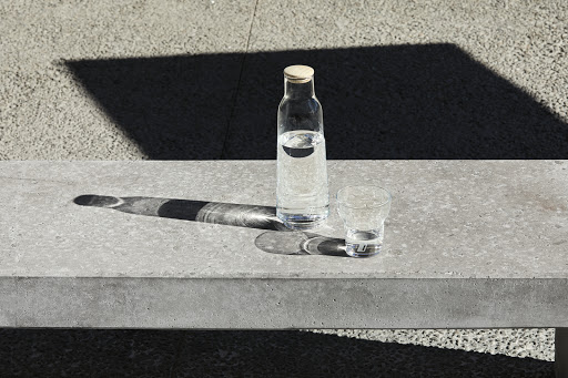 A clear glass water bottle with a cork stopper and a matching glass on a grey concrete bench under bright sunlight.
