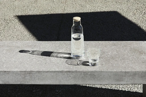 A clear glass water bottle with a cork stopper and a matching glass on a grey concrete bench under bright sunlight.