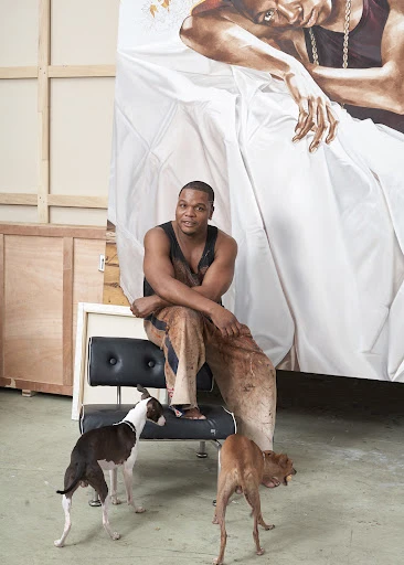 African American man sits on a black bench with two dogs in an art studio in front of his large painting of a reclining person.