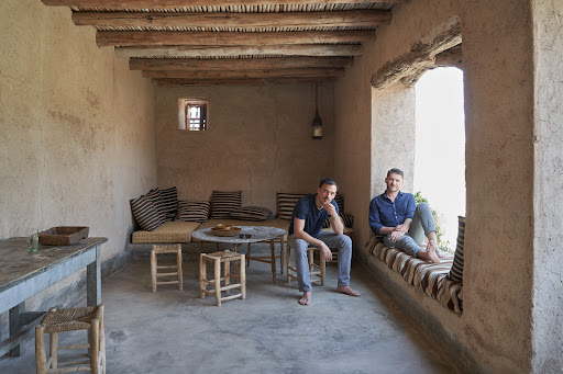 Two men sitting inside a room with light plaster walls, wooden beams, straw furniture, and a window offering views of the outdoors.