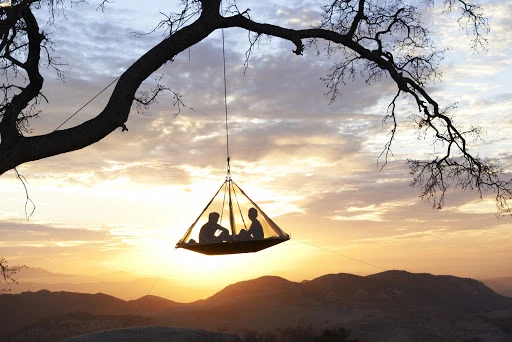 Two people relax in a suspended tent hanging from a tree branch silhouetted against a mountain range and a golden sunrise with cloudy sky.