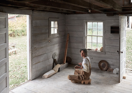 Woman wearing brown overalls sitting cross-legged on a wooden stool inside a rustic whitewashed wooden cabin.