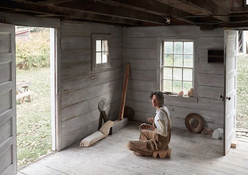 Woman wearing brown overalls sitting cross-legged on a wooden stool inside a rustic whitewashed wooden cabin.