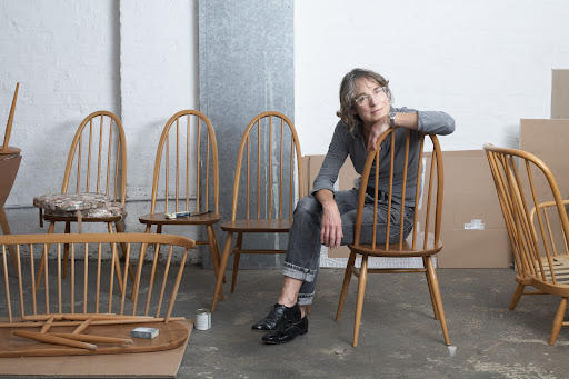Adult woman sits on wooden chair surrounded by disassembled chairs and cardboard boxes in a sparsely furnished room with a concrete floor.