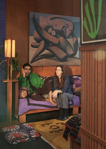 A man and a woman are sitting on a patterned sofa in a wood-paneled room with paintings on the wall. The floor has a patterned rug.