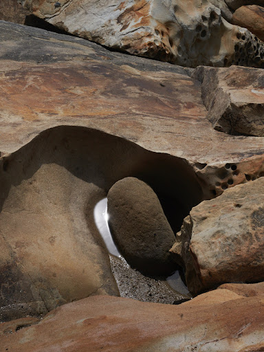 Erosion carved into a rock face, forming a pocket where a smooth river rock rests with sunlight glinting in the water.