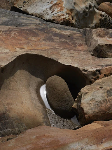Erosion carved into a rock face, forming a pocket where a smooth river rock rests with sunlight glinting in the water.