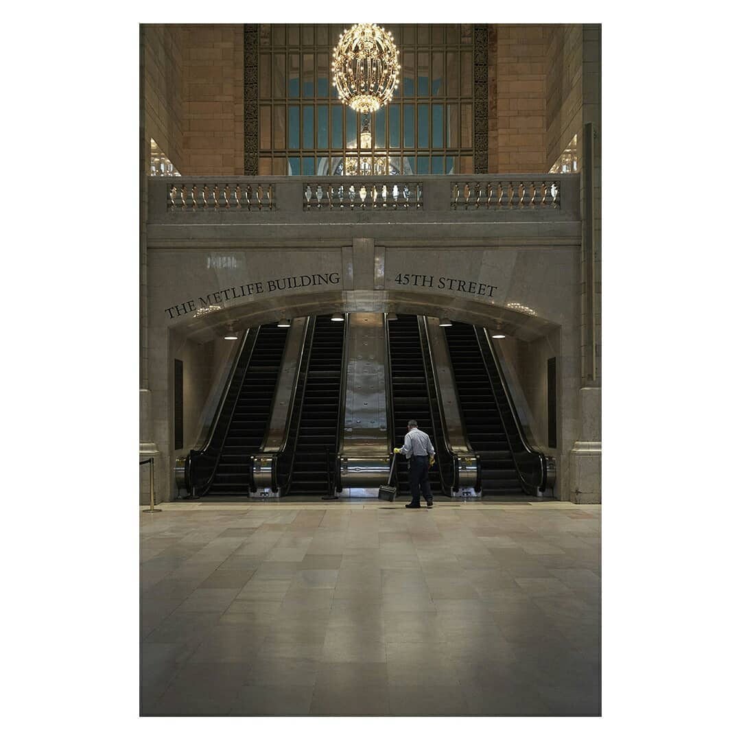 Interior view of a train station with escalators, featuring a maintenance worker and architectural details, including a chandelier and signage.