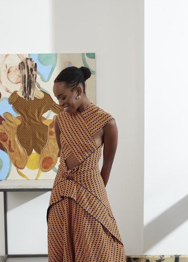 Woman in printed crossover dress smiles slightly with a painting in the background indoors in a gallery with natural lighting.