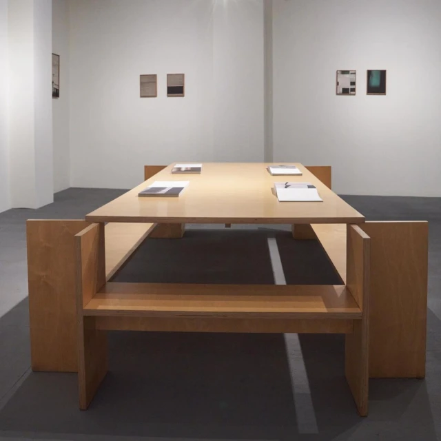 Minimalist art installation featuring a light wood table and bench in a white room, with abstract paintings adorning the walls.