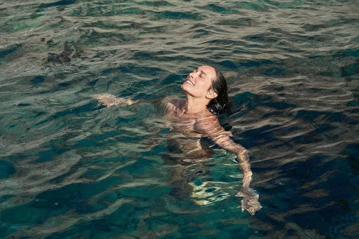 Woman floats in shimmering turquoise ocean water, eyes closed, enjoying a sunny swim, creating a serene and peaceful image of relaxation.