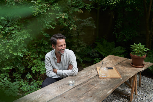 A man with crossed arms sits at a rustic table in a lush outdoor setting, next to a glass of water and a tray with decanter.