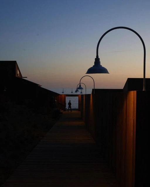Person at end of wooden walkway toward ocean, lamps on sides, twilight sky, creating a sense of depth and tranquility.