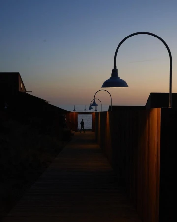 Person at end of wooden walkway toward ocean, lamps on sides, twilight sky, creating a sense of depth and tranquility.