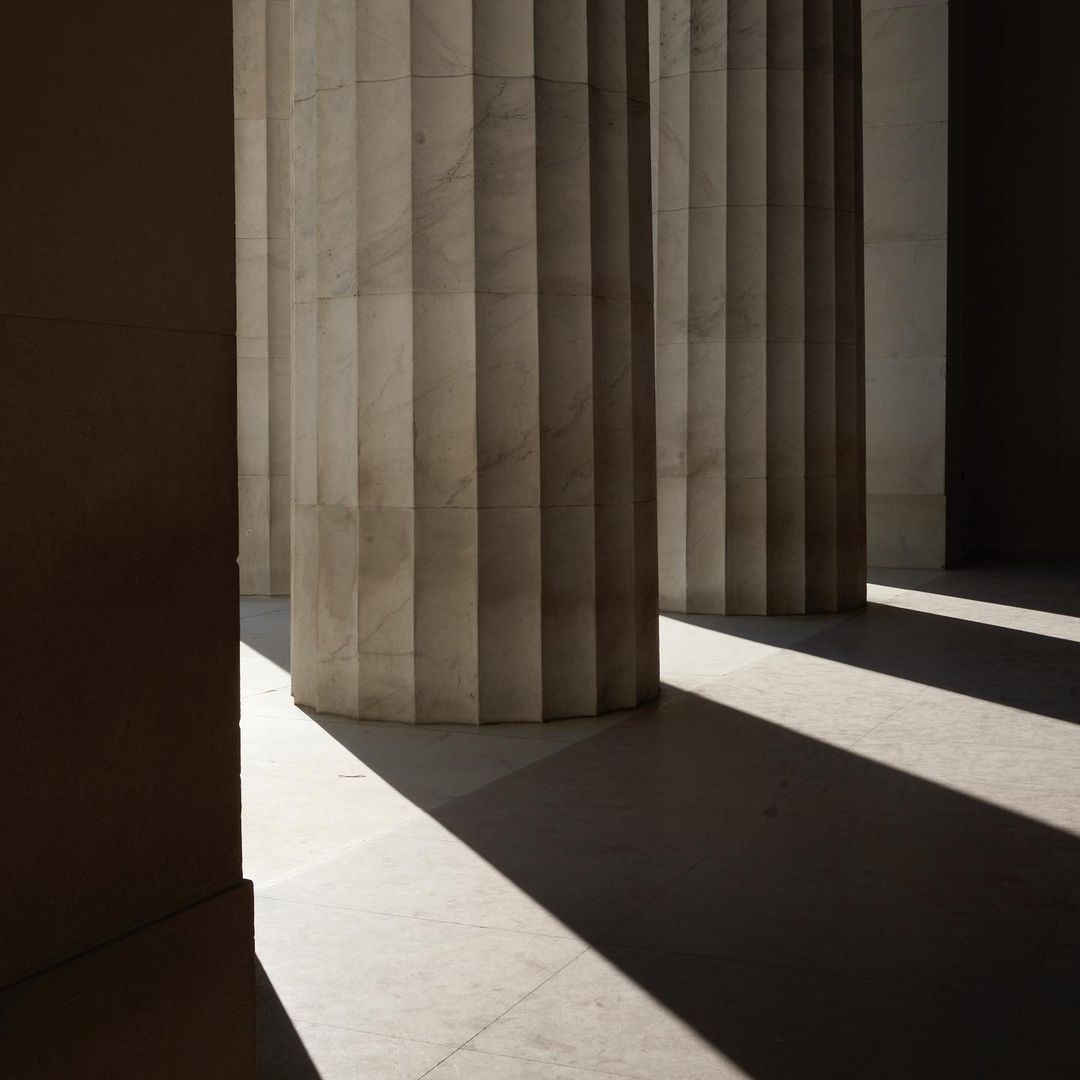 Architectural columns cast long shadows on a light-colored stone floor in an indoor setting, creating a contrast of light and dark.