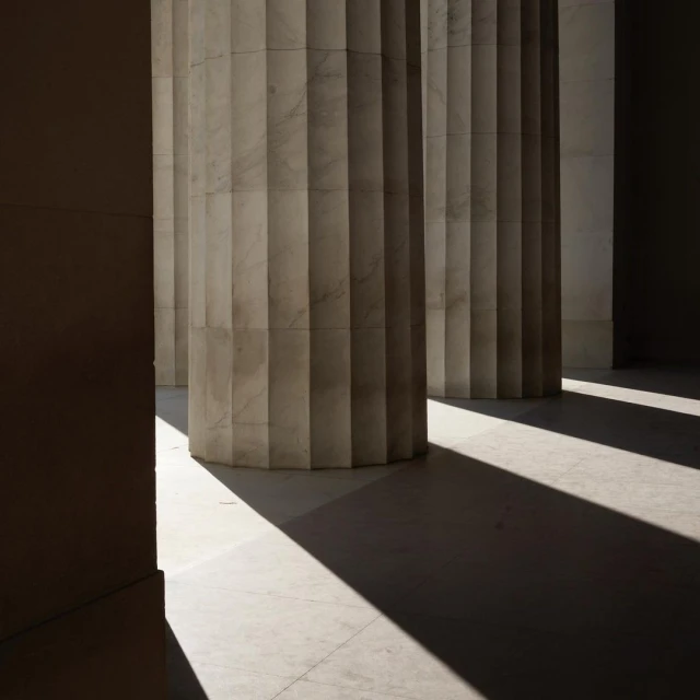 Architectural columns cast long shadows on a light-colored stone floor in an indoor setting, creating a contrast of light and dark.