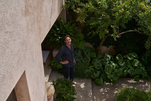 Eye-level shot of an adult male in a dark outfit standing in a courtyard filled with greenery near a building on a sunny day.