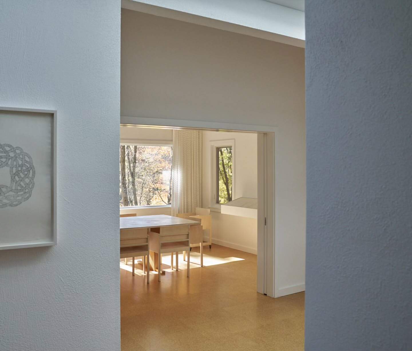 Interior view of an art-filled dining room, featuring a simple wooden table and chairs, light wood flooring, and minimalist white walls.