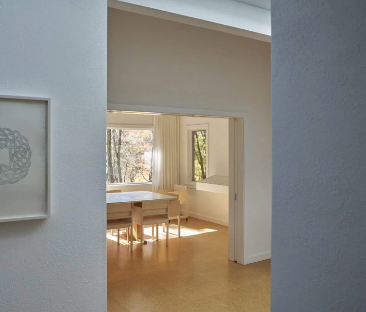 Interior view of an art-filled dining room, featuring a simple wooden table and chairs, light wood flooring, and minimalist white walls.