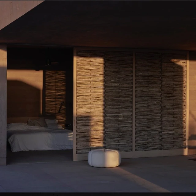 Modern bedroom interior with a bed visible through a sliding door and a white ottoman on a concrete floor illuminated by golden sunlight.