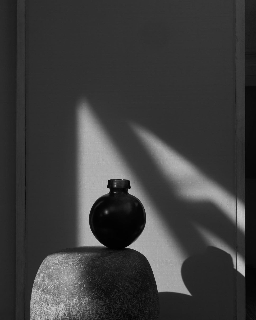 Monochrome photo of a dark, round vase on a rough, textured surface, set against a wall with a striking triangular light pattern.