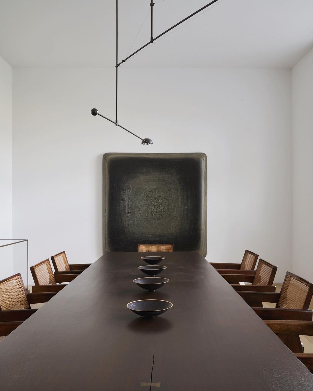 Interior photo of a dining room featuring a long dark wood table set with black bowls, wooden chairs, a modern light fixture, and art.