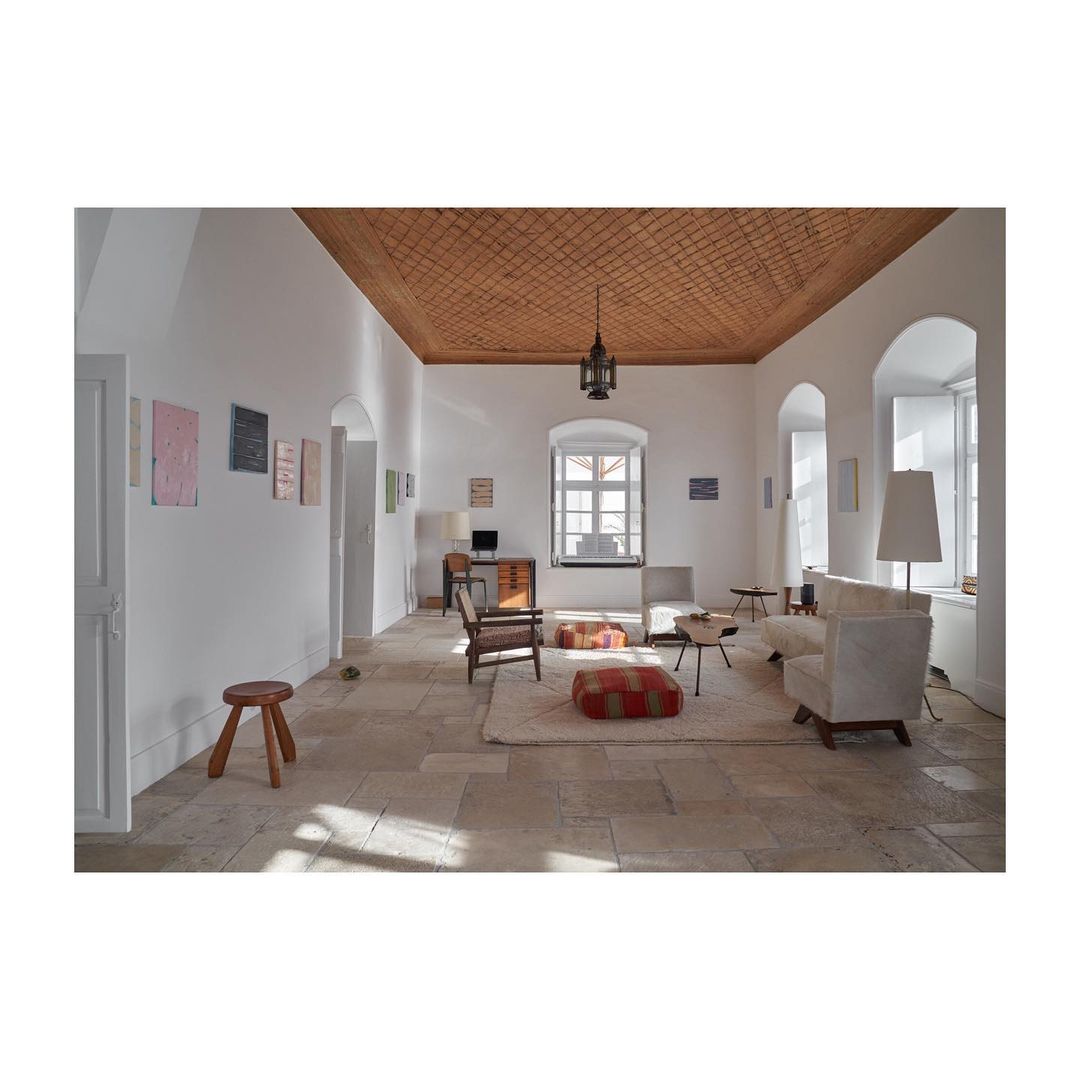 Bright, sunlit living room with light-beige stone floors, neutral furniture, and wood-latticed ceiling featuring eclectic art and design.