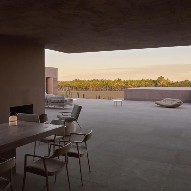 Outdoor patio with a concrete table and chairs under a covered area, overlooking green vegetation at sunset. Minimalist interior design.