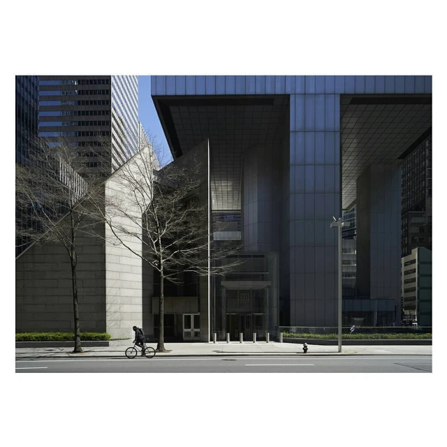 Cyclist rides past modern architecture in an urban environment on a sunny day. Tall buildings are in the background.