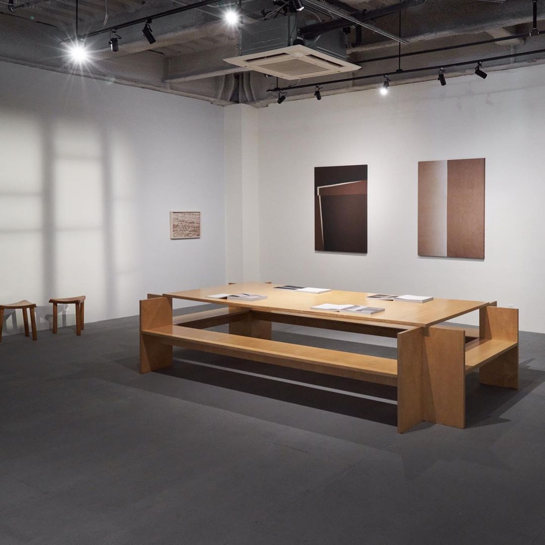 Minimalist art gallery featuring a light wood table with documents, wooden stools, and abstract art pieces on white walls.