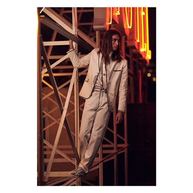Adult male fashion model posing in off-white suit with black piping, climbing metal structure at night with blurred neon lights in background.