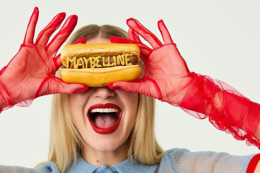 Woman holding a hotdog with 'Maybelline' spelled in mustard, wearing sheer red gloves, bright red lipstick, and a light blue sweater.