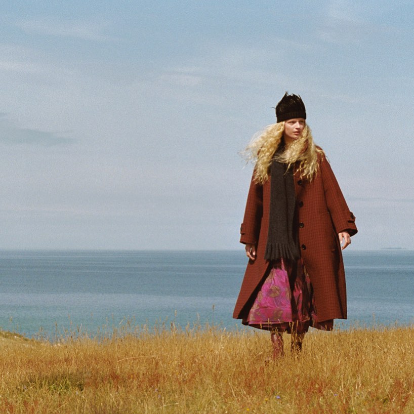 Woman with long blonde hair wearing an overcoat and black hat stands in a field with the sea and sky in the background.