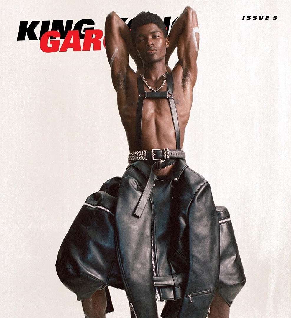 Shirtless Black man in leather harness poses with arms behind head, draped with a black leather jacket against a white background.