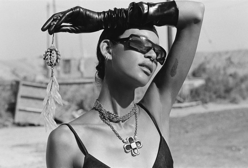 Portrait of a woman with sunglasses and an earring in her hand, wearing a black dress and jewelry, set against an outdoor backdrop.