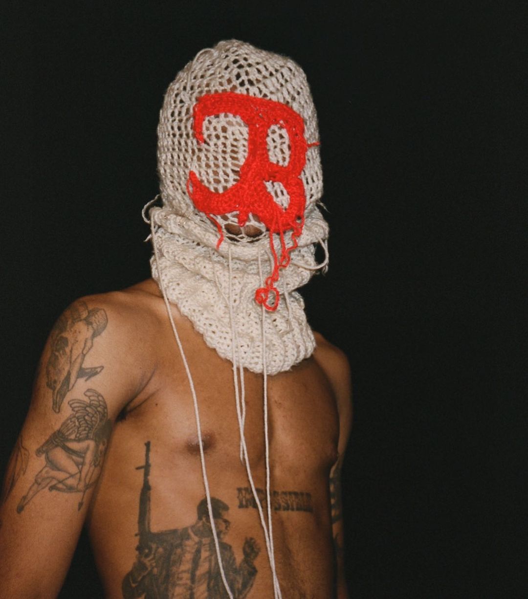 A shirtless African American man with tattoos wearing a balaclava with a red letter "B" against a dark background in a studio shot.