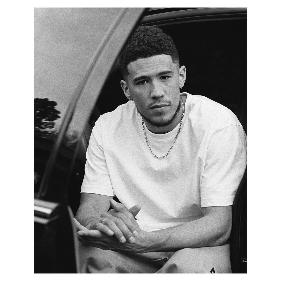 Monochrome portrait of Devin Booker wearing a white t-shirt and chain necklace seated inside a vehicle