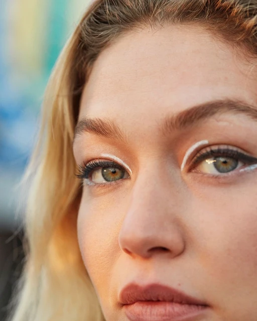 Close-up portrait of a woman with blond hair, wearing distinctive white eyeliner, and a neutral facial expression.