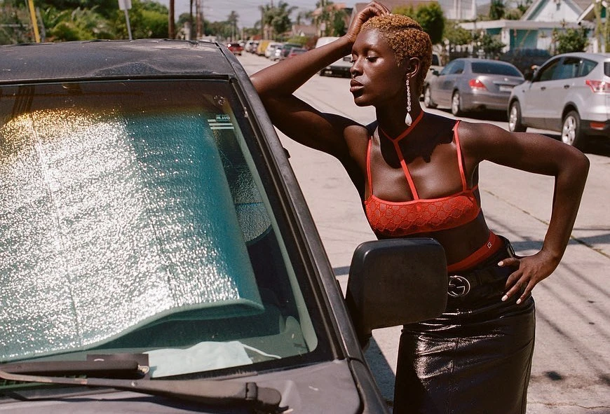 An African American adult female in a red lingerie top and a black patent leather skirt poses next to a dark car on a sunny street.