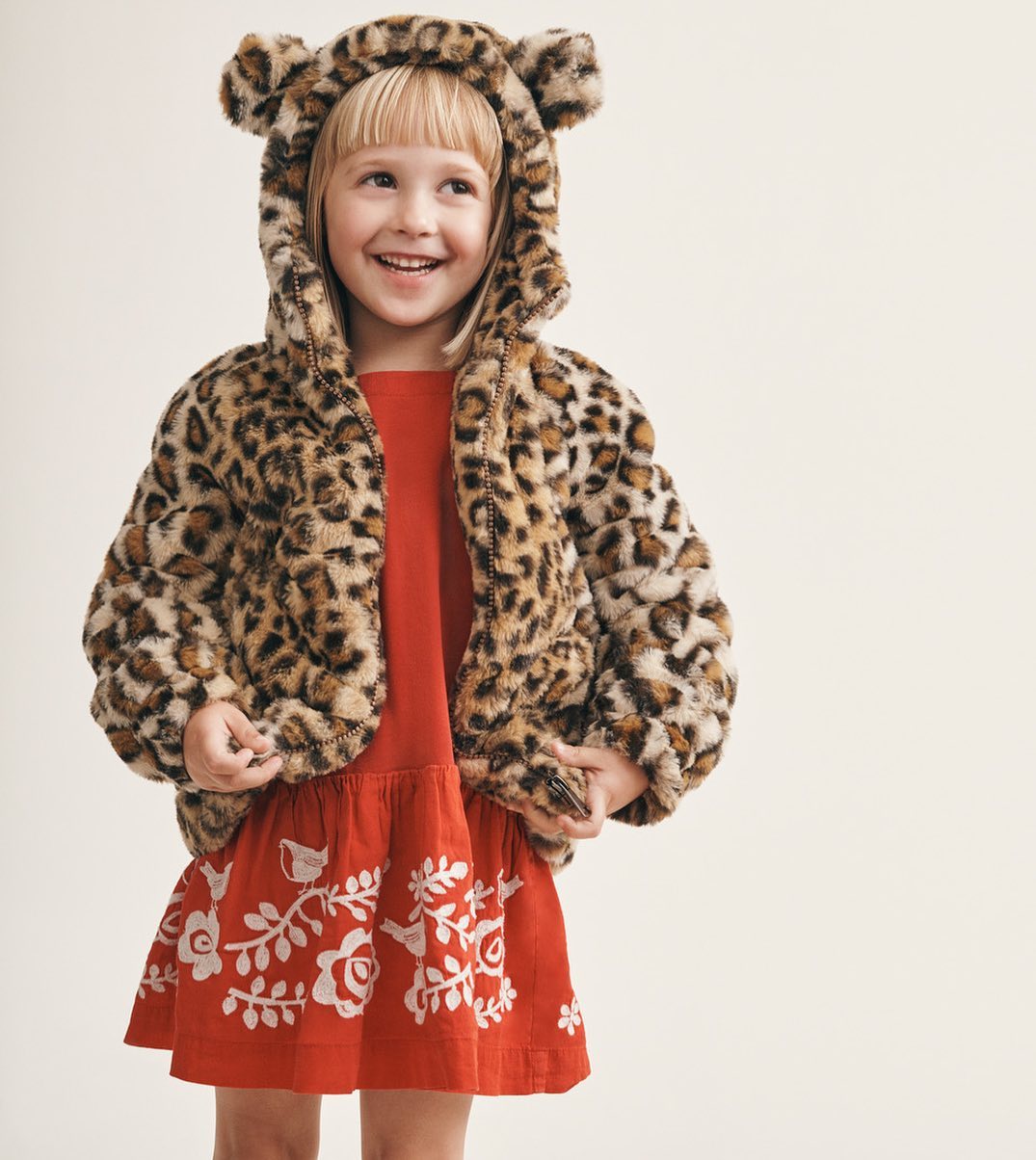 A young girl smiles wearing a red dress with floral embroidery and a hooded faux fur coat with bear ears against a light backdrop.