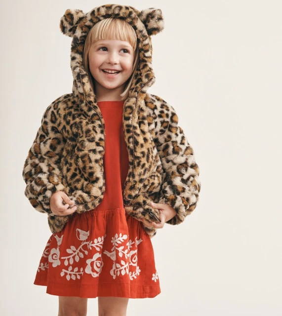 A young girl smiles wearing a red dress with floral embroidery and a hooded faux fur coat with bear ears against a light backdrop.