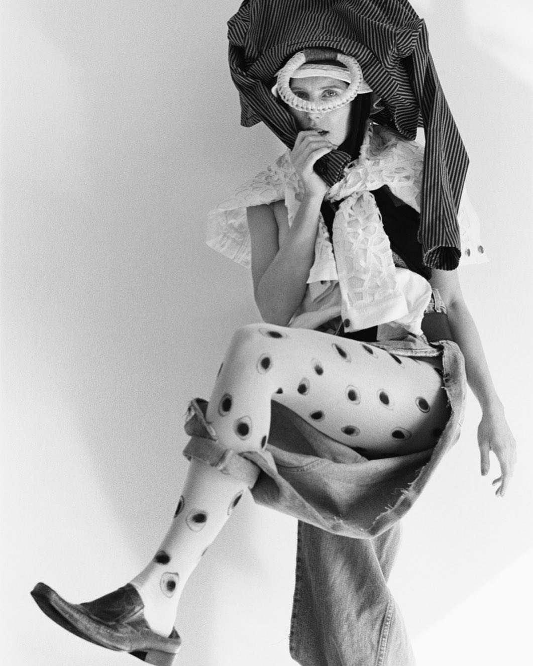Avant-garde fashion portrait featuring a person in a unique ensemble with patterned tights, oversized clothing, and a quirky headpiece.