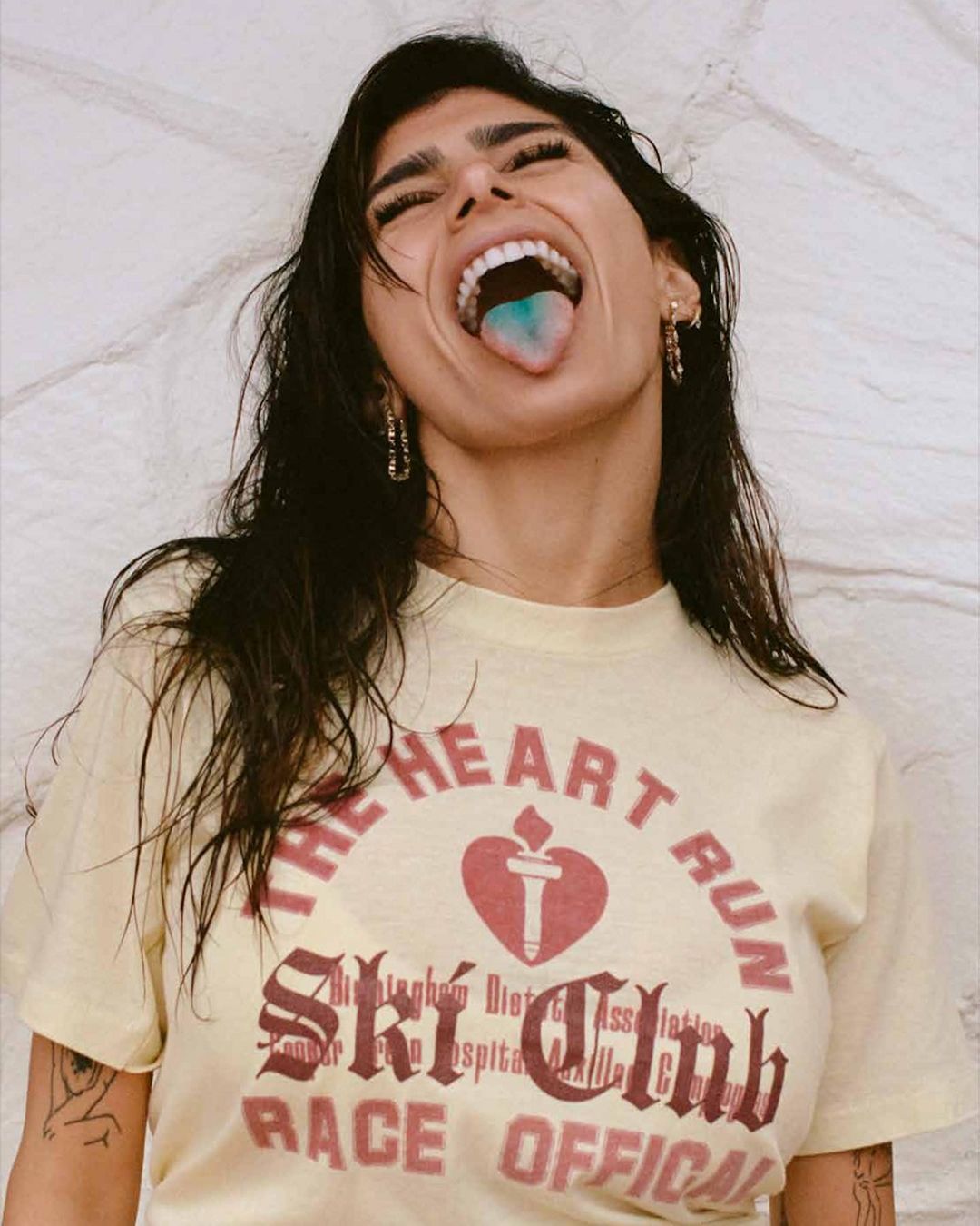 Young woman with open mouth and blue tongue shows off vintage t-shirt with heart design. Indoor shot against textured wall.