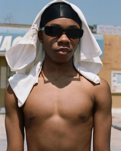 Close-up of a shirtless young adult male wearing sunglasses, a black durag, and a white towel draped over his head against an outdoor background.