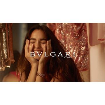 Smiling woman with hands on face in Bulgari jewelry advertisement. Gold bracelet visible, dress behind. Studio lighting, medium shot.
