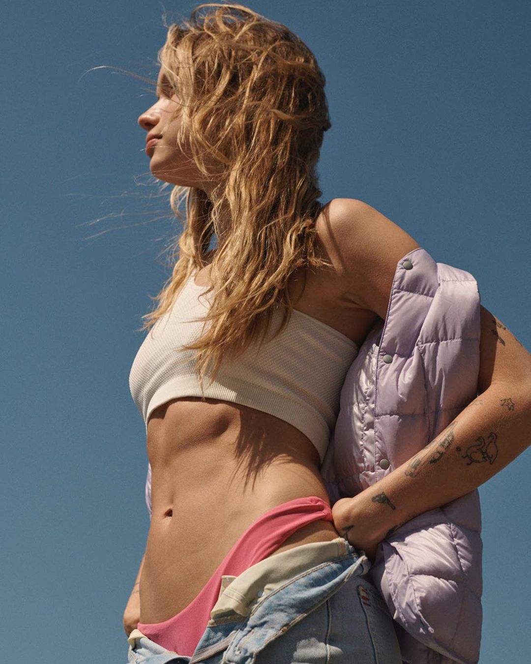 Blonde woman gazes into the distance, wearing a white tank top, pink bikini bottoms, and denim overalls, holding a lavender puffer jacket.