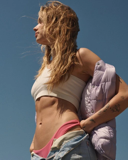 Blonde woman gazes into the distance, wearing a white tank top, pink bikini bottoms, and denim overalls, holding a lavender puffer jacket.
