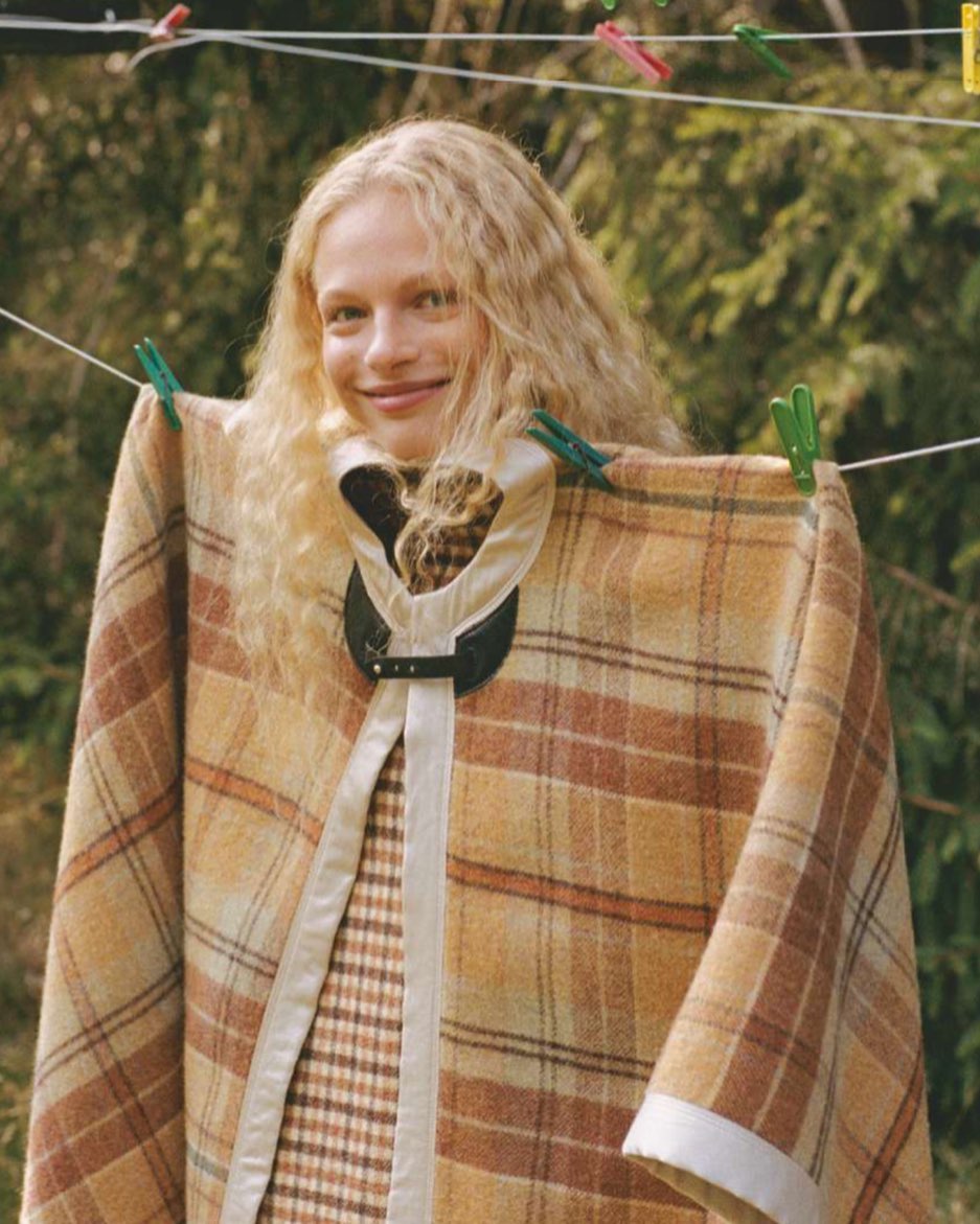 Smiling blond woman in a plaid shawl with a black clasp is hanging on a clothesline in a sunny, rural, outdoor setting.